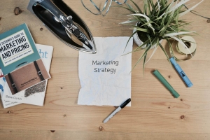 Stack of marketing books, plants, and crumpled paper reading "Marketing Strategy" sit on a large wooden desk